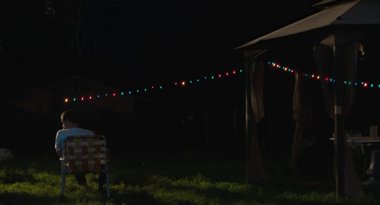 Test Pattern (2019) movie still: wide — A dark grassy backyard at night illuminated by a string of multicolored lights. A lone man sits in a folding…