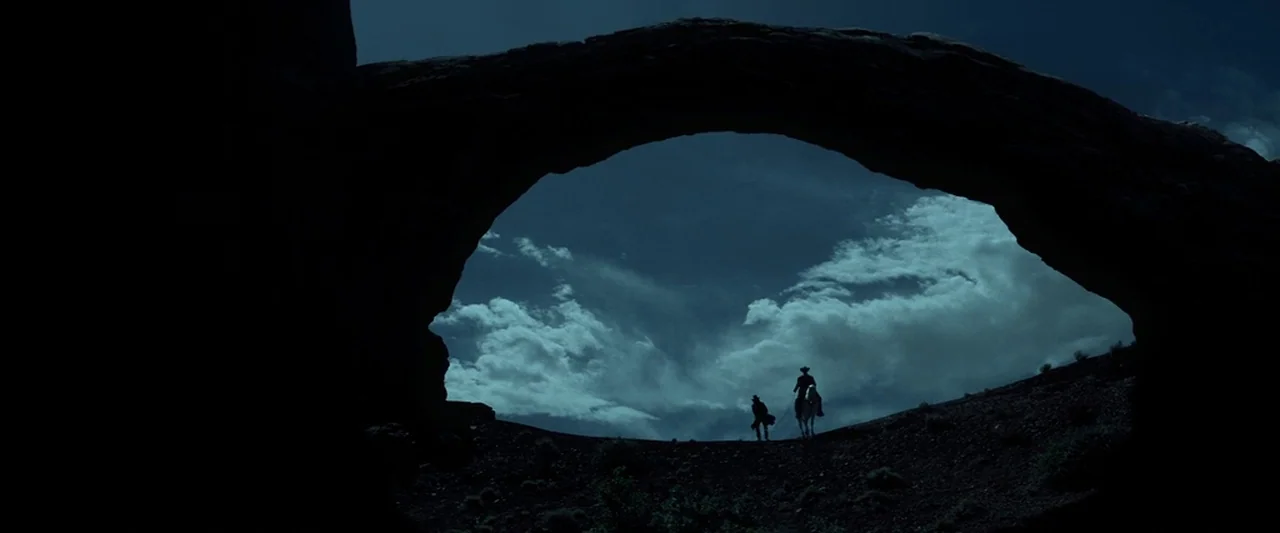 The Lone Ranger (2013) movie still: wide — A dark natural stone arch frames a view of a cloudy sky above a hill. Two riders on horseback move across…