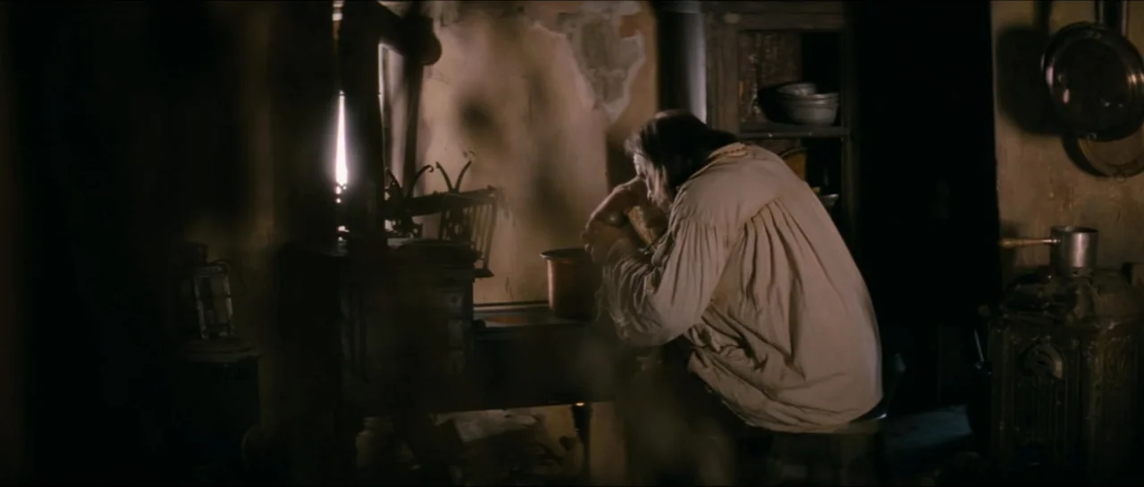 A Farewell to Fools (2013) movie still: medium — A dimly lit rustic wooden interior with aged walls and a cast iron stove. A man in a simple shirt…