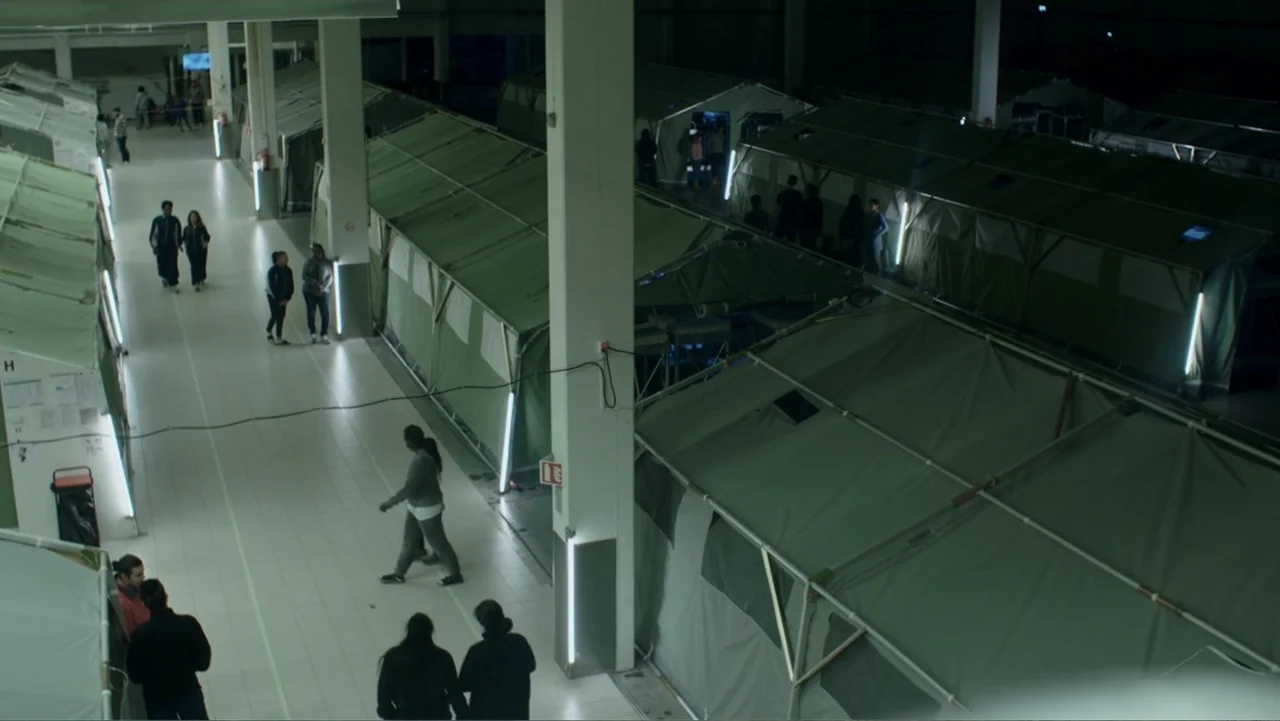 Code of Trust (2019) movie still: wide — A large indoor warehouse space is filled with rows of green canvas camping tents. Multiple individuals walk…