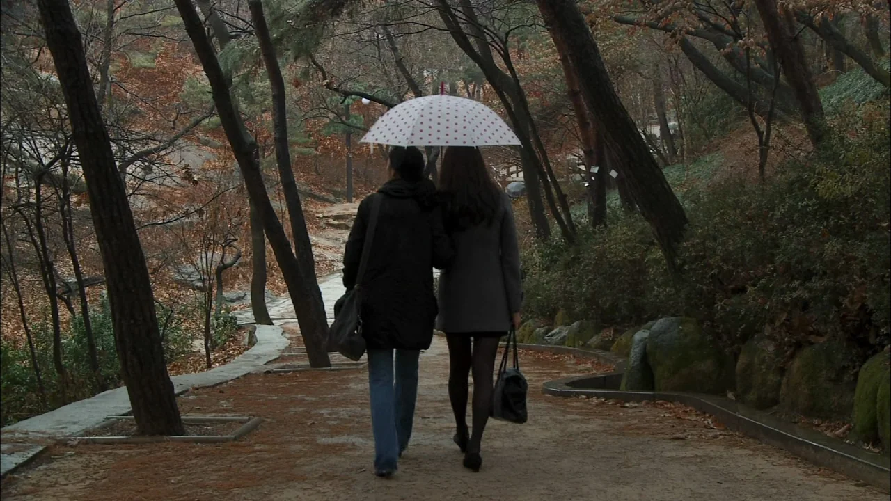 Oki’s Movie (2010) movie still: fullbody — A winding dirt path snakes through a wooded area with bare, winter trees. Two women walk away from the…