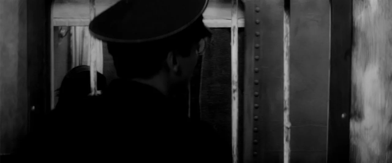 Europa (1991) movie still: medium — A dimly lit industrial environment features heavy vertical iron bars separating two figures. A man in a peaked…