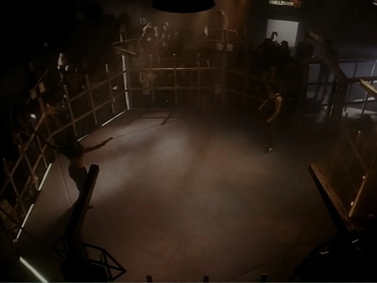 Firepower (1979) movie still: wide — A dark, industrial-style fighting arena features a central cage enclosed by metal fencing. Two male fighters…
