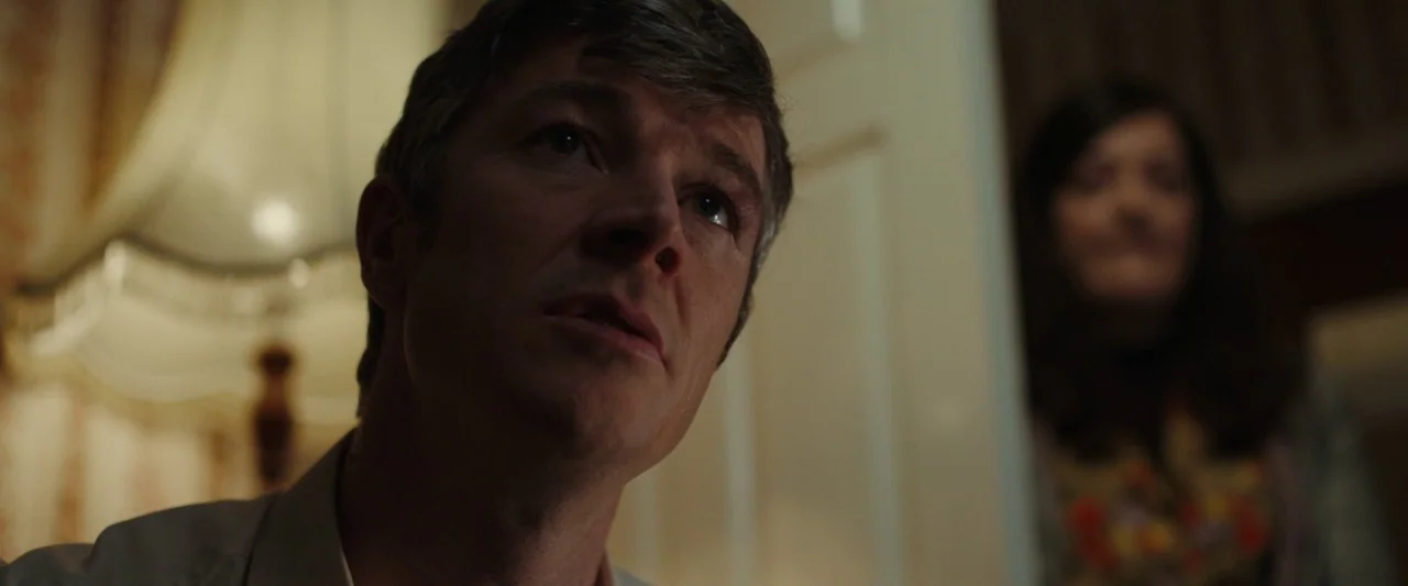 Extra Ordinary (2019) movie still: closeup — A dimly lit residential hallway at night features a man in the foreground. A woman stands obscured by…