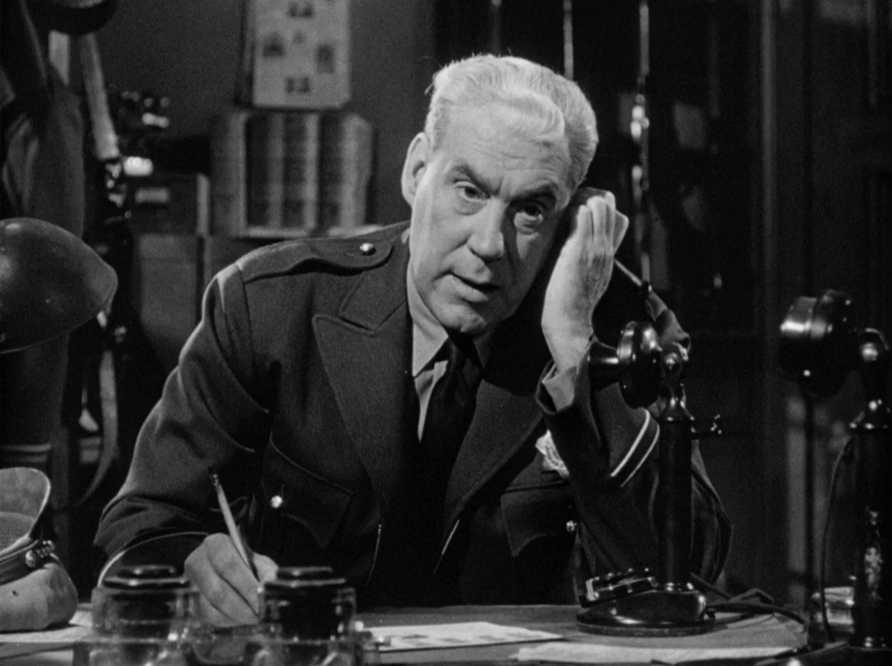 Gun Crazy (1950) movie still: medium — A dimly lit room with stacks of files and various vintage desk items. A man in uniform sits at a desk while…