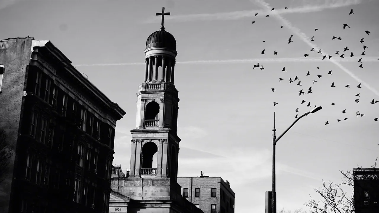 Kokomo City (2023) movie still: wide — A stone church tower rises beside a dark multi-story brick apartment building. A large flock of birds circles…