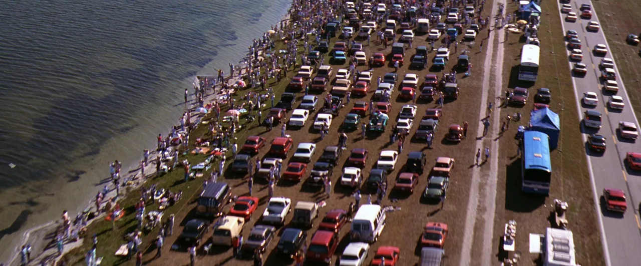 Contact (1997) movie still: establishing — A vast field packed with numerous parked cars stretches along the shore of a lake. People walk between…
