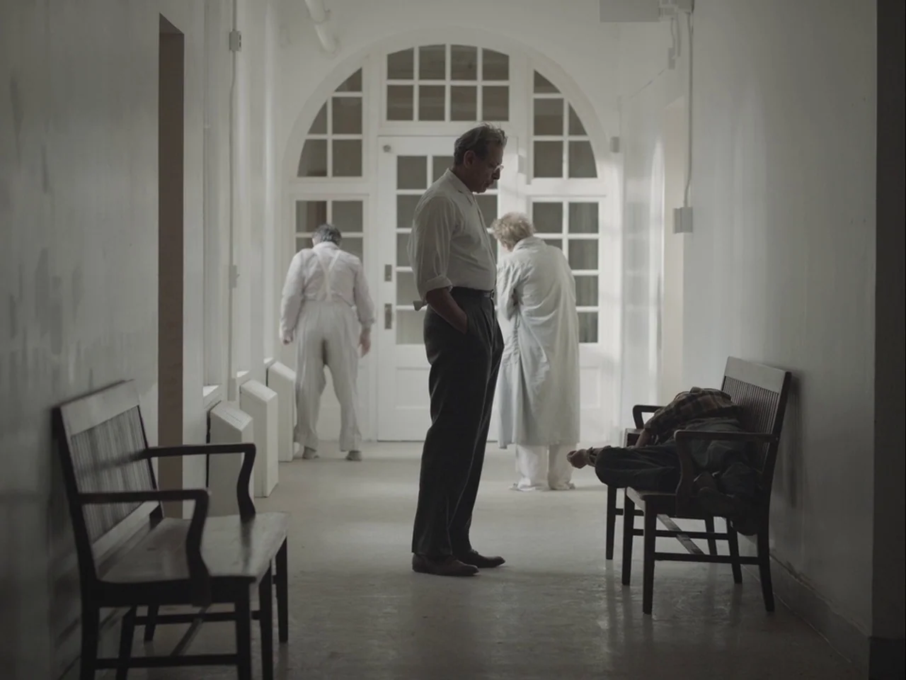 The Mountain (2018) movie still: fullbody — A long, white, sterile hallway features an arched glass door at the far end. Several individuals occupy…