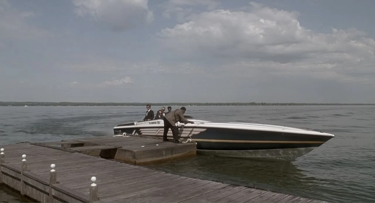 F/X2 (1991) movie still: wide — A wooden pier extends into a vast body of water under a cloudy sky. One man secures a sleek racing powerboat to the…