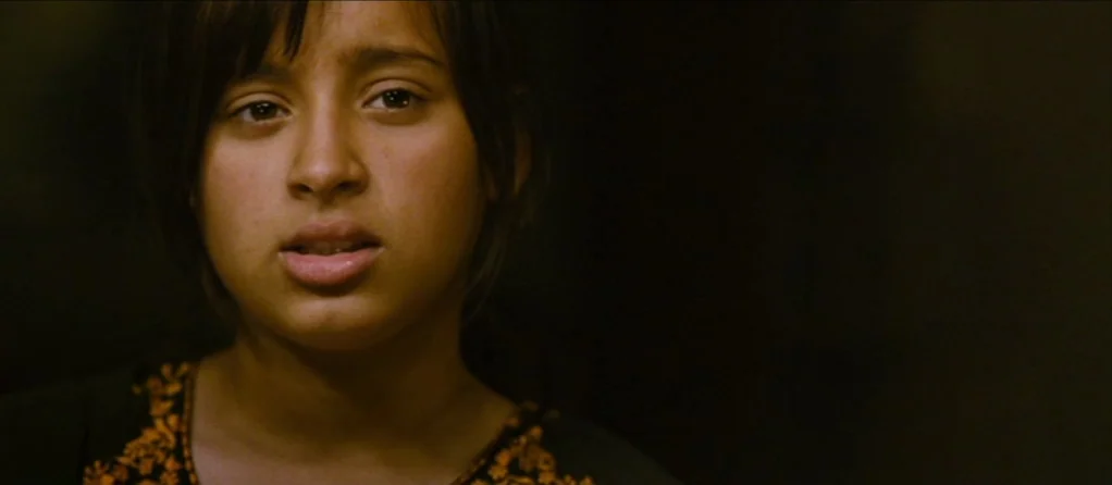 Brick Lane (2007) movie still: closeup — A dimly lit room with a dark, indistinct background. A young girl faces forward with an attentive expression.
