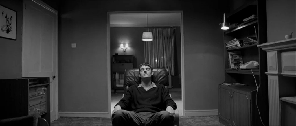 Control movie still: medium — A sparsely furnished 1990s living room features a dark leather armchair positioned centrally. A young man sits in the…