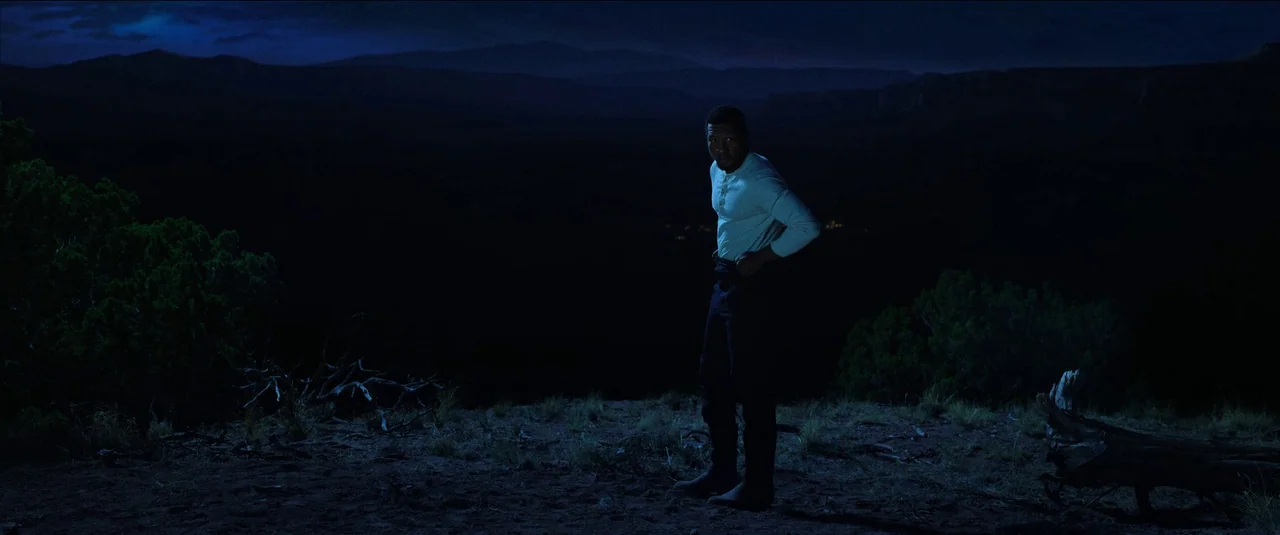 The Harder They Fall (2021) movie still: fullbody — A man stands alone on a desolate canyon edge under a dark night sky. He turns his head toward…