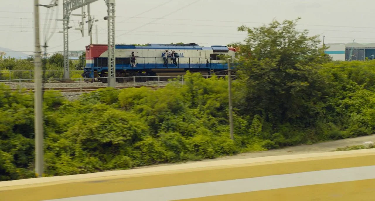 Train To Busan movie still: wide — A diesel locomotive with blue, white, and red livery moves across elevated railroad tracks. Dense green…