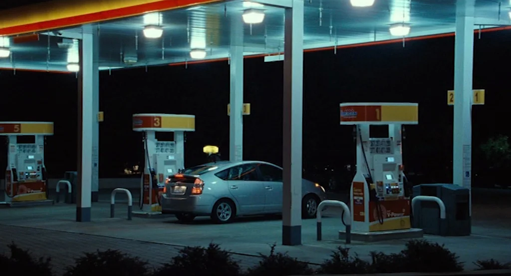Louder Than Bombs (2015) movie still: wide — A night view of a modern gas station forecourt under bright fluorescent canopy lighting. A silver sedan…
