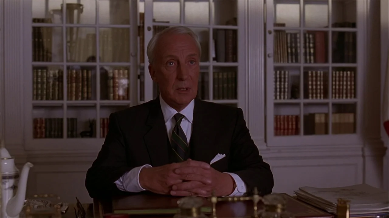M. Butterfly (1993) movie still: medium — An interior space features white bookshelves filled with books behind a wooden desk. A man in a suit sits…