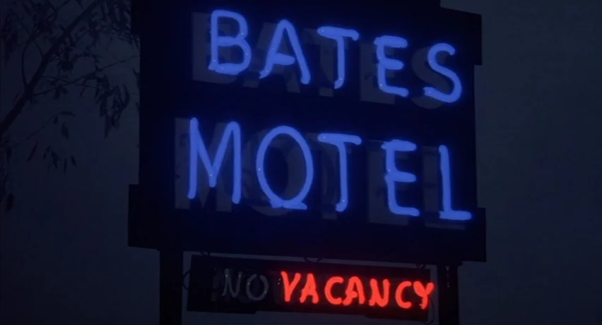 Psycho 2 (1983) movie still: closeup — A dark outdoor setting features a black rectangular sign with glowing blue lettering. Below the primary sign,…