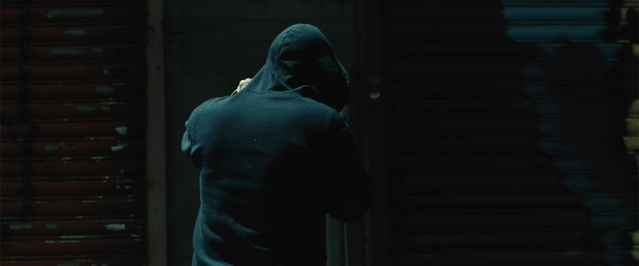 Skin (2009) movie still: medium — A dark night exterior features a metal security gate on a storefront wall. A solitary person wearing a dark hoodie…