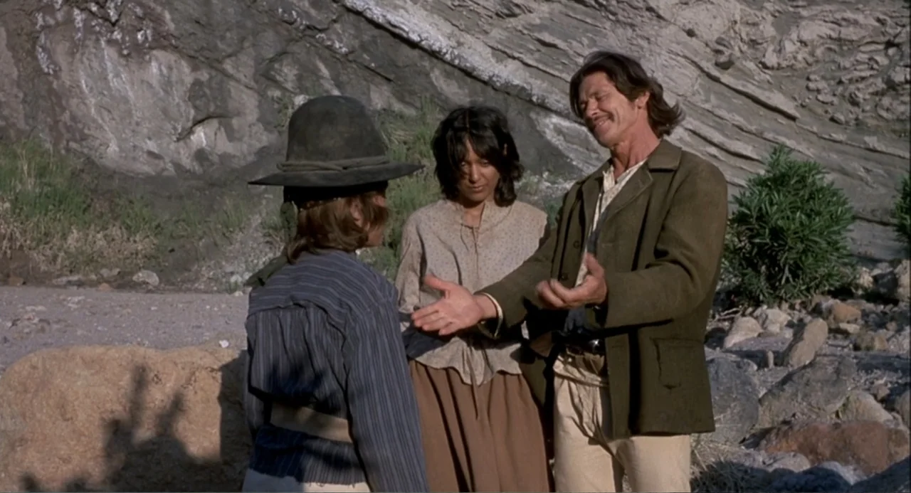 Chato's Land (1972) movie still: medium — A rocky, arid outdoor environment features a large stone cliff face background. An older man gestures…