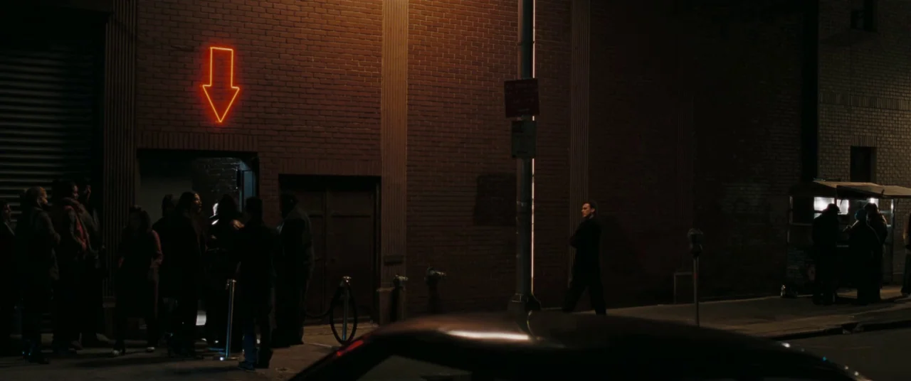 Two Lovers (2008) movie still: wide — A brick building at night features a glowing neon down arrow above an entrance. A group of people waits in…