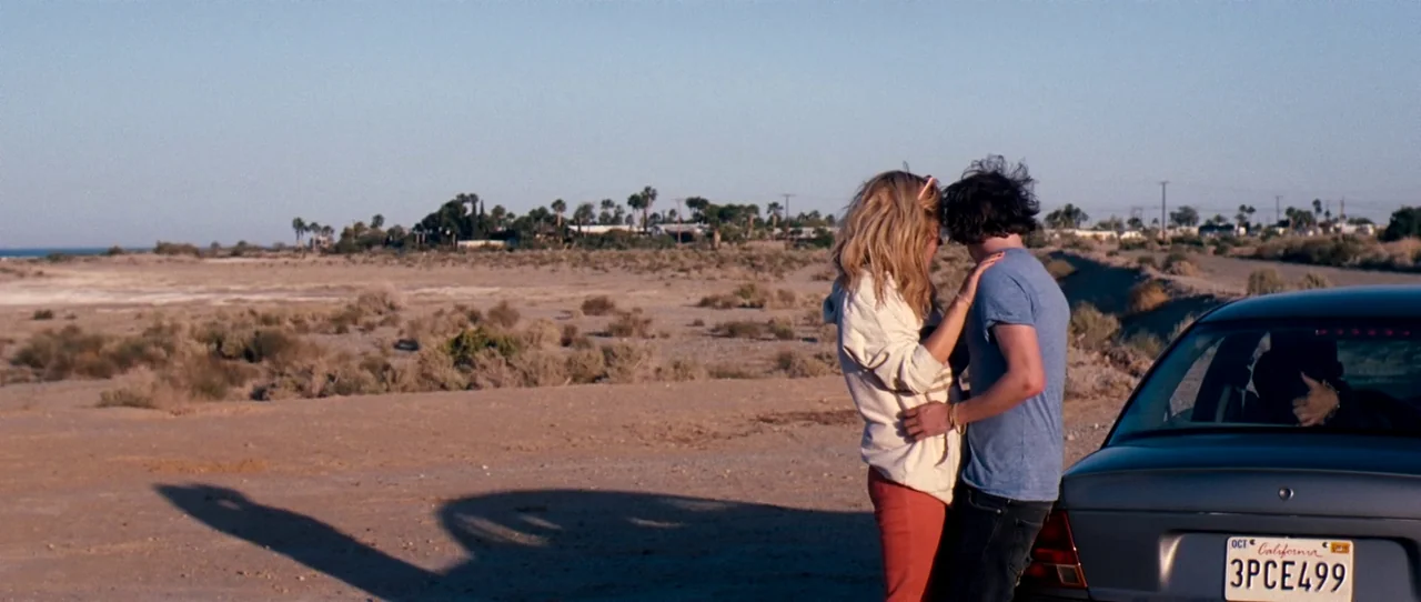 Little Birds (2011) movie still: wide — A dry, flat desert landscape under a clear sky with scrubby vegetation. A young couple stands against the…