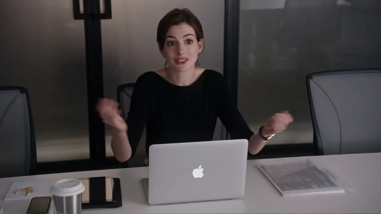 The Intern (2015) movie still: medium — A modern office meeting room with white tables and dark glass partitions. A woman in a black shirt sits at…
