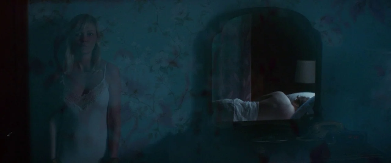 Woodshock (2017) movie still: medium — A dimly lit room features vintage floral wallpaper and a large wall mirror. A woman in a white slip dress…