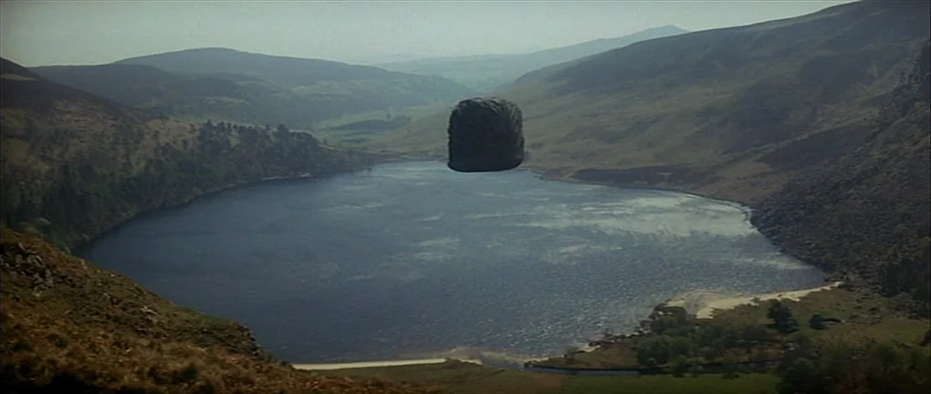 Zardoz (1974) movie still: establishing — A vast, deep blue lake is nestled between rolling, grass-covered hills and steep slopes. A lone…