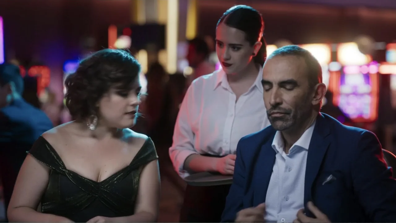 Lecciones para canallas (2022) movie still: medium — A casino floor filled with blurred colorful neon gaming machines in the background. Two…