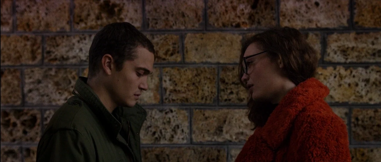 Love (2015) movie still: medium — A porous stone wall serves as the backdrop for two people standing outside. The man and woman face each other in a…