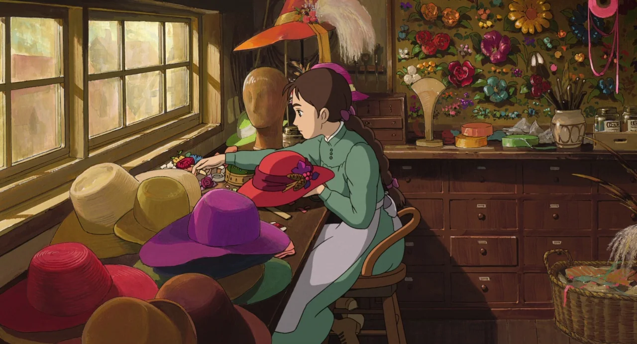 Howl’s Moving Castle (2004) movie still: medium — A cluttered workshop is filled with various hats and floral wall decorations. A young woman sits…