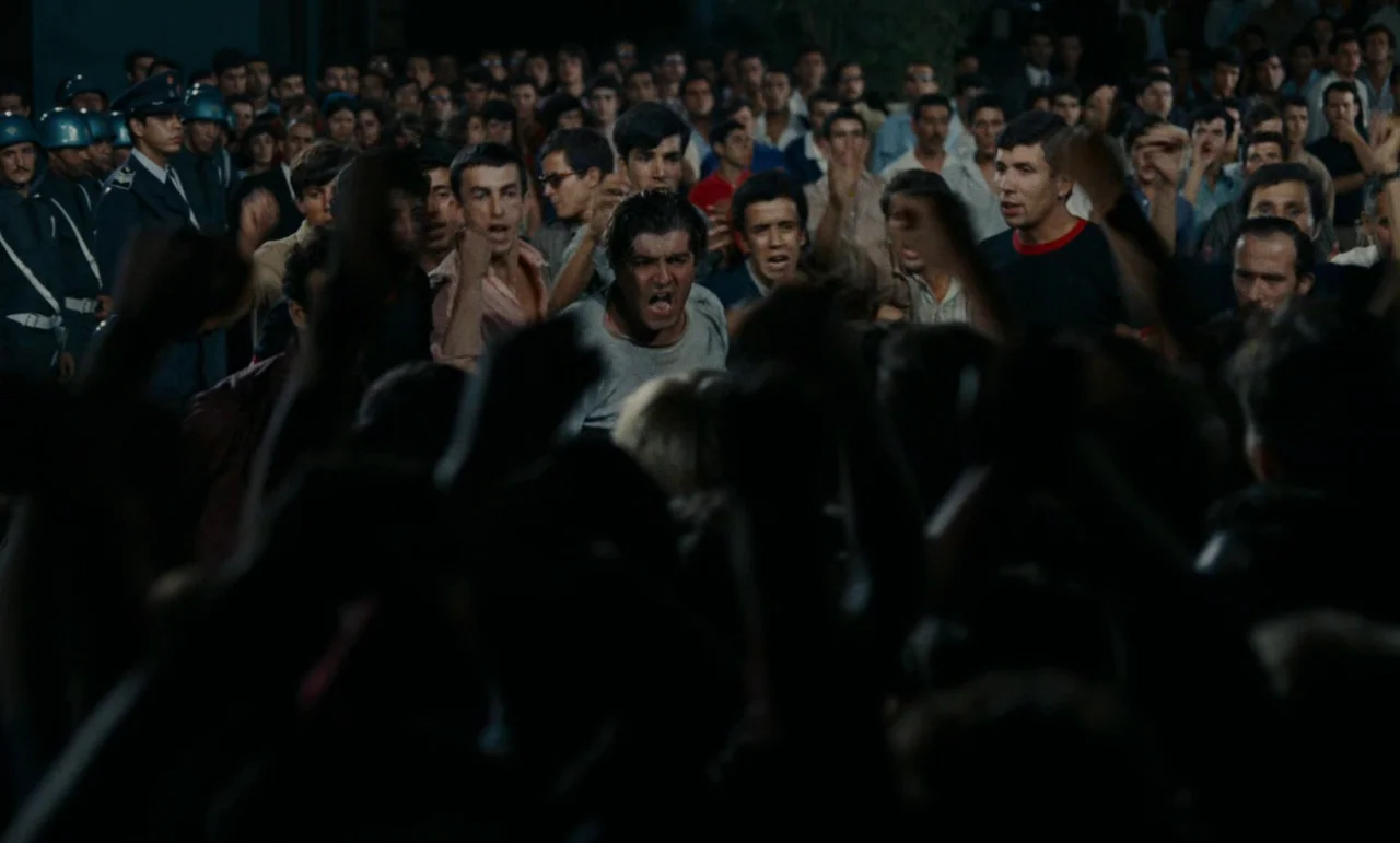 Z (1969) movie still: medium — A dense group of men gathers at night during a public protest. The central subject stands in the middle of the crowd…