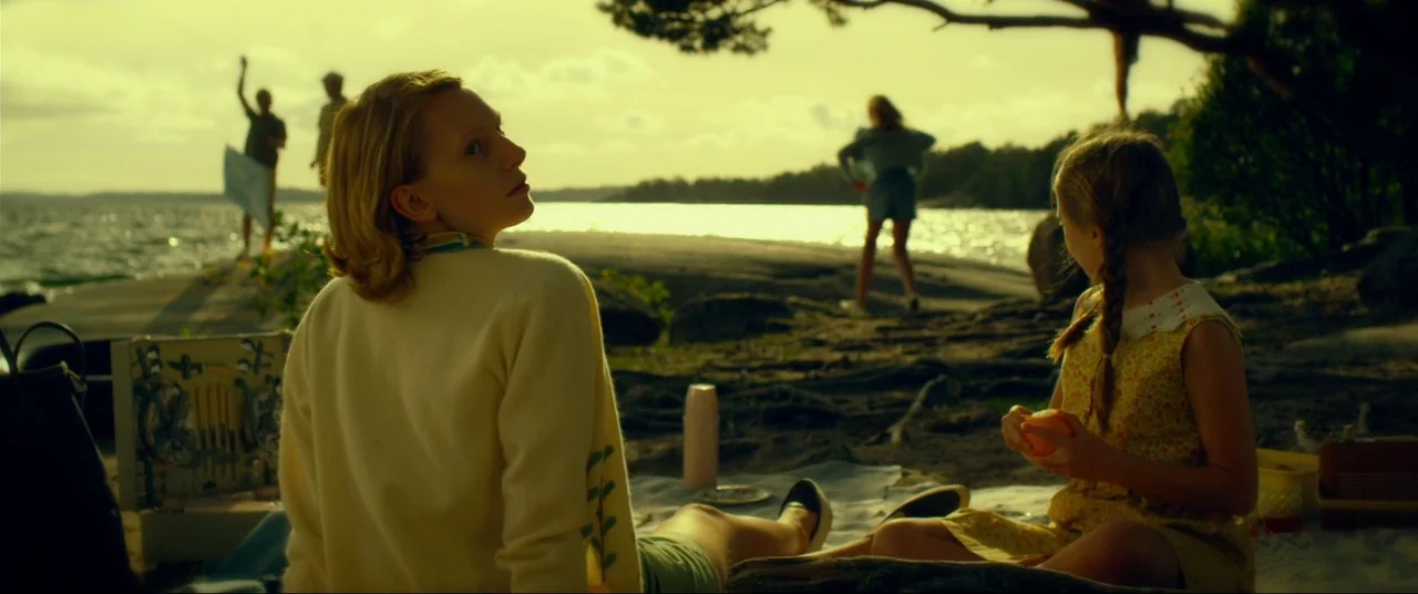 The Girl With The Dragon Tattoo (2011) movie still: medium — A woman and young girl sit on a blanket near a bright shoreline. Other people stand in…