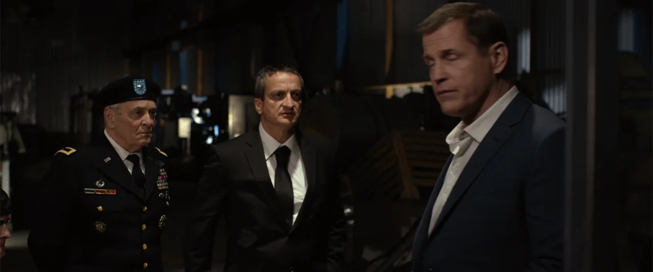 The Condemned 2 (2015) movie still: medium — Three men stand inside a dimly lit industrial space with mechanical equipment in background. An older…