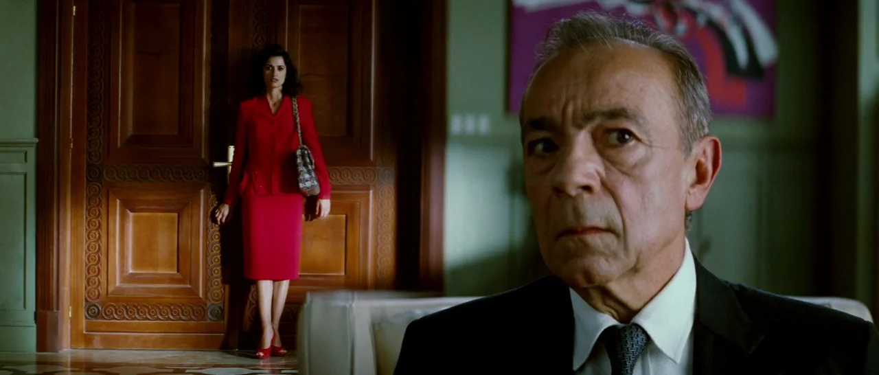 Broken Embraces (2009) movie still: medium — A formal interior with sage-colored paneled walls and a large, carved wooden door. An older man in the…