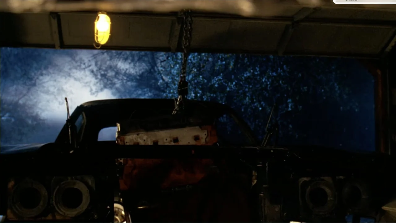 Chain Letter (2010) movie still: wide — A dark residential garage at night contains an open vehicle chassis with its engine removed. A heavy metal…
