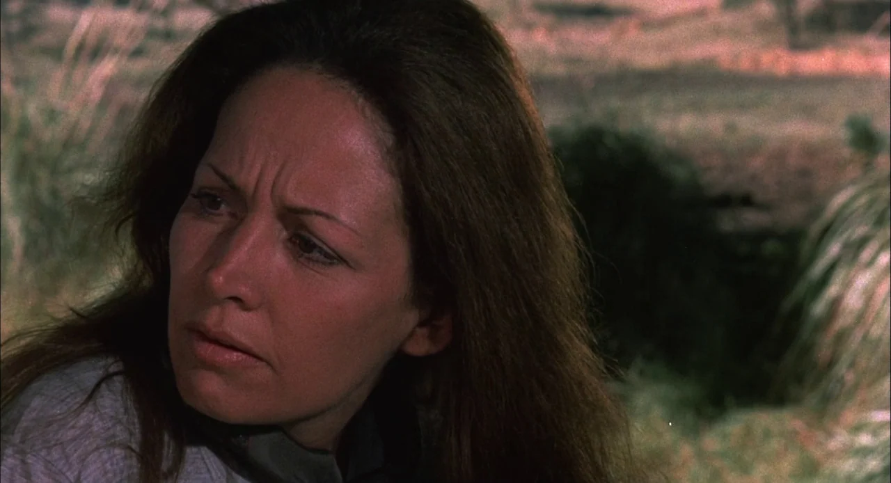 Bring Me The Head of Alfredo Garcia (1974) movie still: closeup — A woman stands in a dry, grassy outdoor environment under natural sunlight. She…