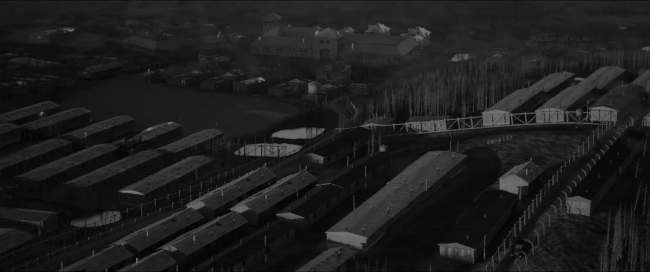 Bau, Artist at War (2025) movie still: establishing — An aerial view displays multiple long wooden barracks arranged in ordered rows at night.…