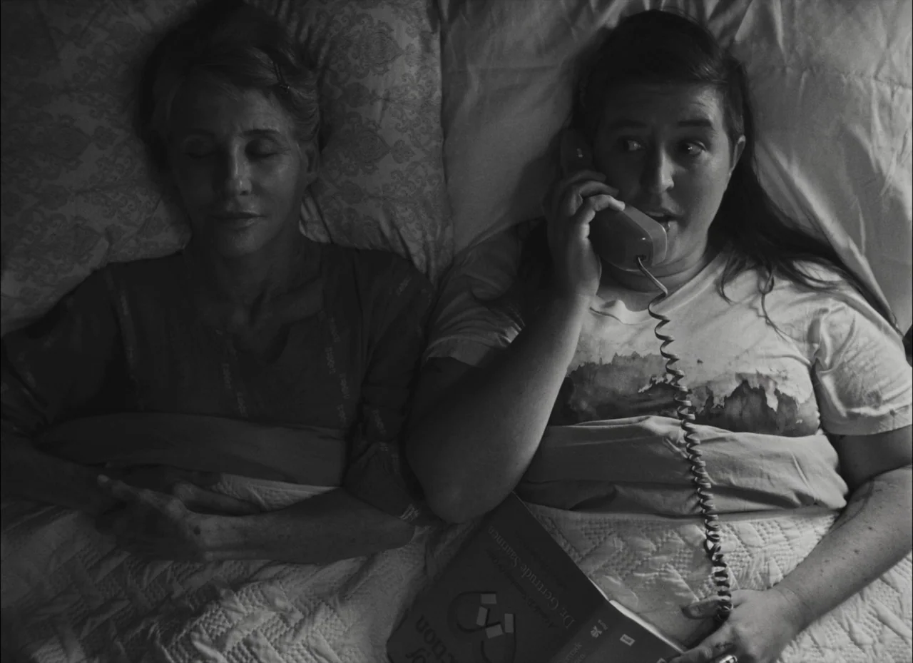 Fremont (2023) movie still: medium — Two women lie side by side on a patterned white quilt in a dark room. One woman is asleep while the other holds…