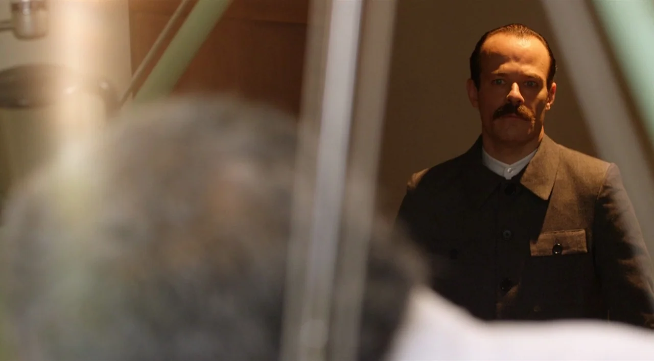 Dance of Reality (2013) movie still: medium — A man in a brown uniform stands in a room with neutral walls. He faces an unseen person located in the…