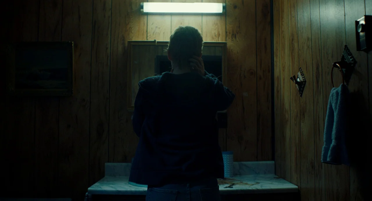 My Animal (2023) movie still: medium — A dark bathroom featuring wood-paneled walls and a marble vanity top with a mirror. A person stands with…