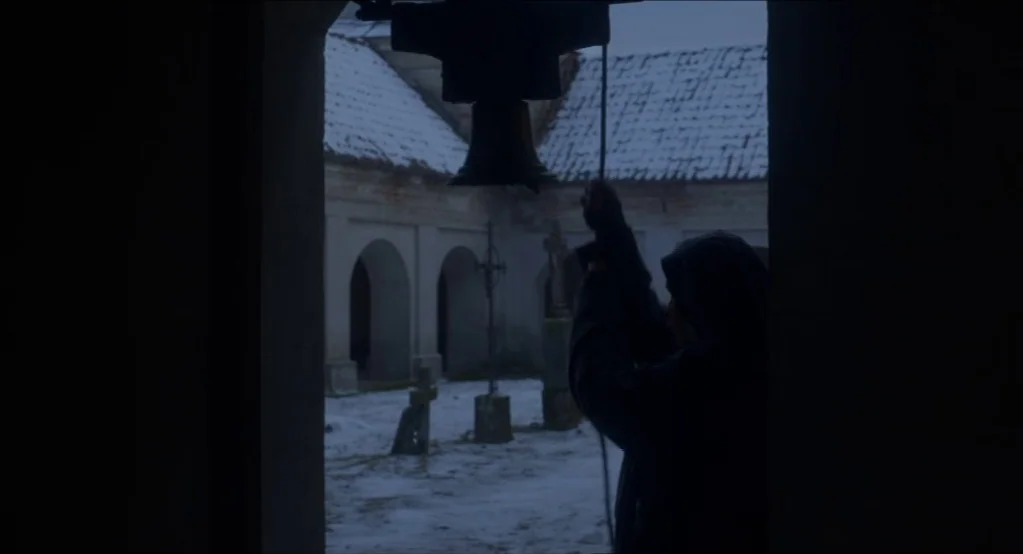 The Innocents (2016) movie still: medium — A dark stone bell tower overlooks a snow-covered courtyard with stone arches and crosses. A person in…