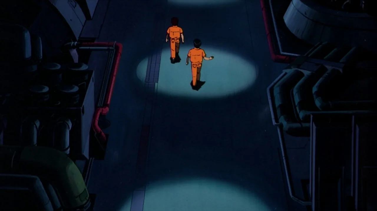 Akira (1988) movie still: wide — An industrial space with metallic piping and a concrete floor under pools of light. Two figures in orange jumpsuits…