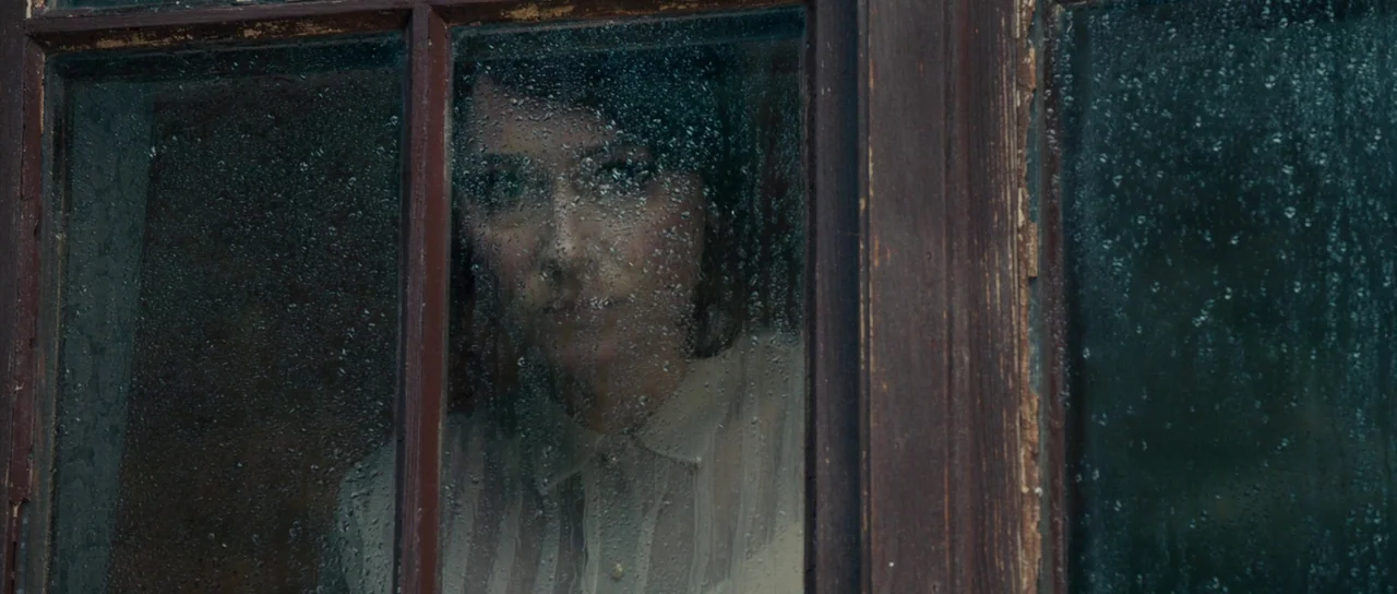 The Duke of Burgundy (2014) movie still: closeup — A woman looks out through a dark-framed wooden window covered in raindrops. The glass pane…