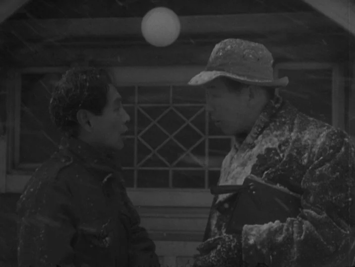 The Idiot (1951) movie still: medium — A residential building exterior during a snowfall features a glowing light above the door. Two men in thick…