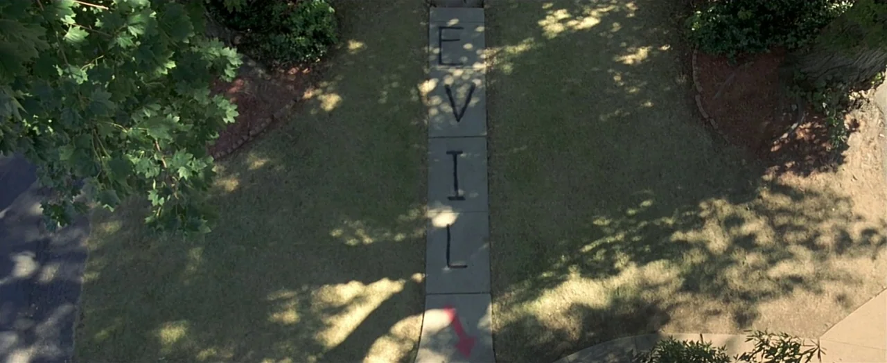 Little Children (2006) movie still: establishing — A concrete sidewalk walkway cuts through a residential lawn with trees overhead. The word EVIL is…