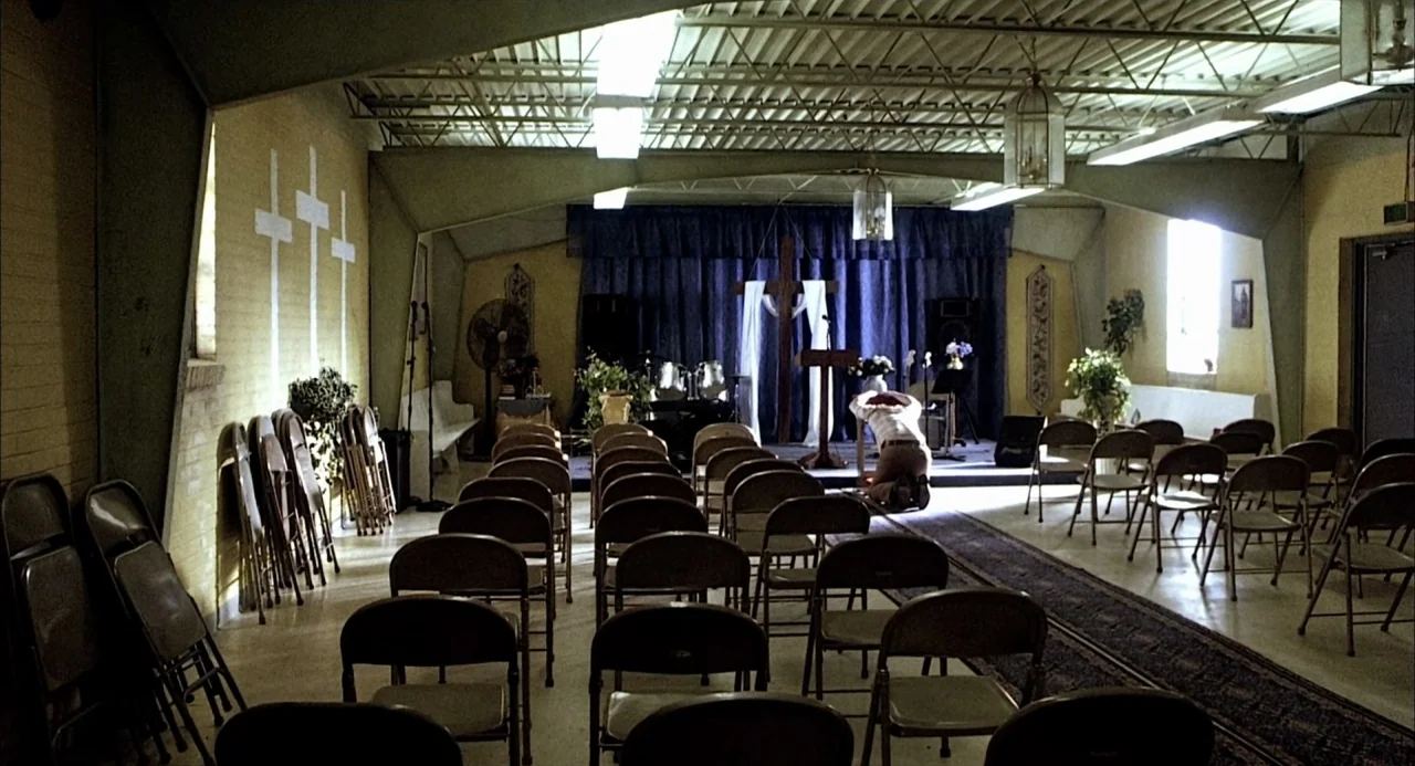 21 Grams (2003) movie still: wide — A spare church hall with rows of metal folding chairs and yellow-painted walls. A lone person kneels in prayer…