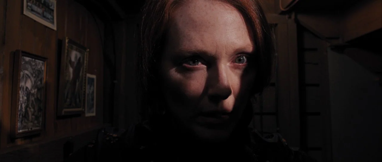 Carrie (2013) movie still: closeup — A dark indoor space features a paneled wooden wall decorated with framed pictures. A woman stares intensely at…