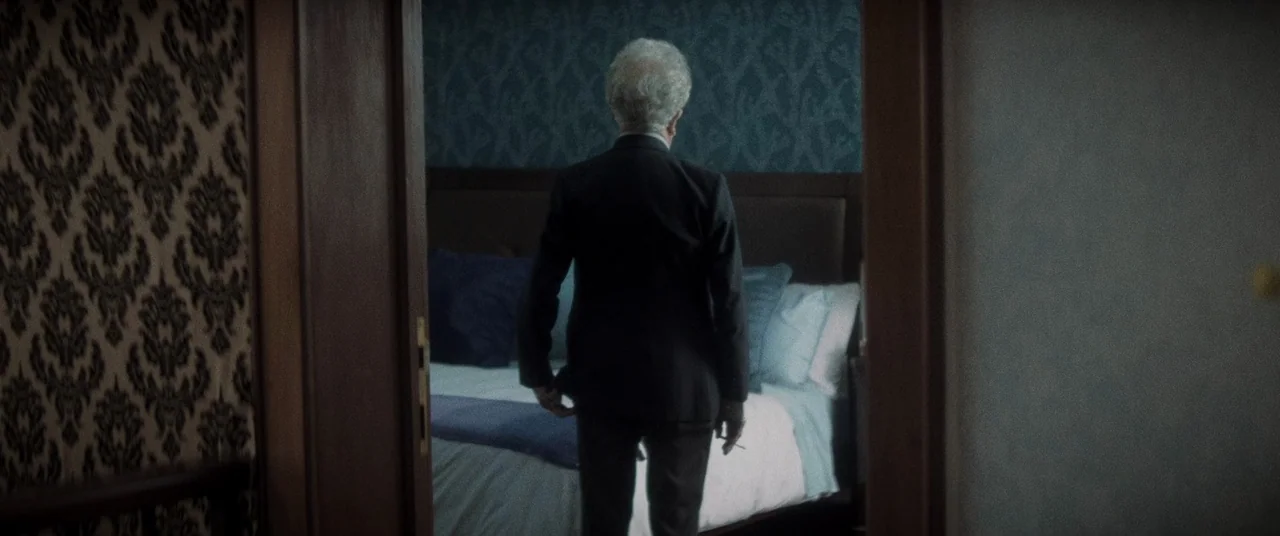 La Llorona (2019) movie still: medium — A dimly lit room with patterned wallpaper features a large unmade bed. An elderly man in a suit stands…