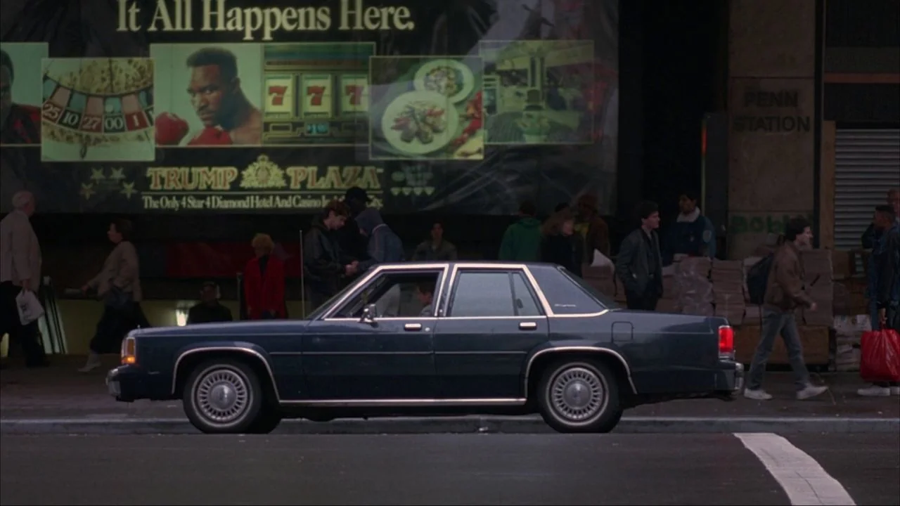 Bad Lieutenant (1992) movie still: wide — A dark sedan drives along a New York City sidewalk in front of Penn Station. A large Trump Plaza billboard…