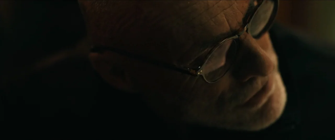 Guns Up (2025) movie still: closeup — A dimly lit interior space with the subject obscured by heavy shadow. A person wearing glasses tilts their…